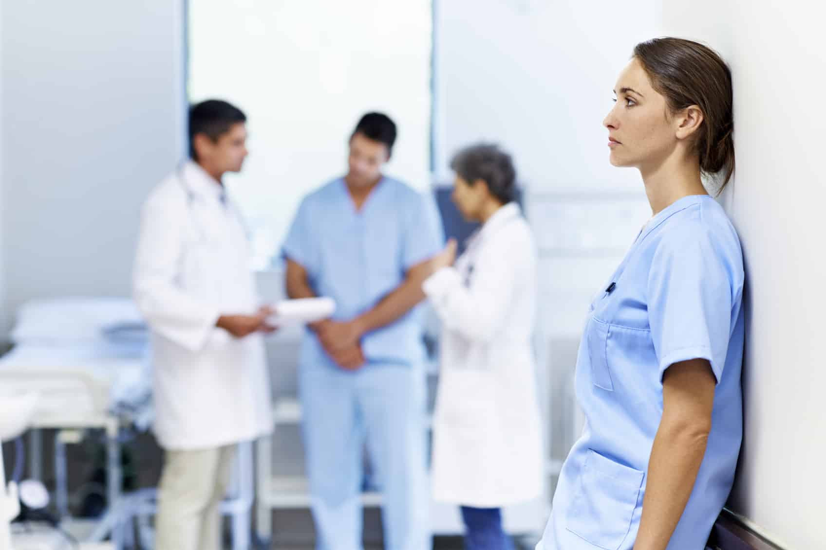 Shot of a tired looking nurse leaning against a wall with colleagues in the background
