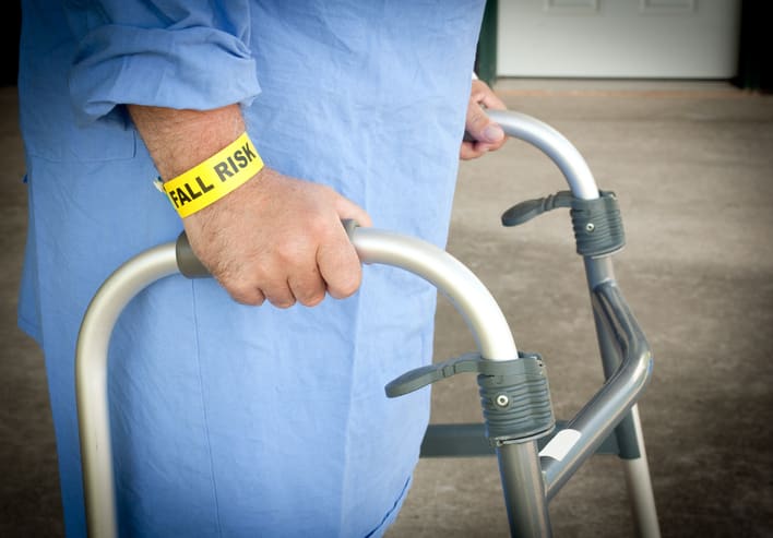A patient walking with a walker wearing a fall risk bracelet and blue gown