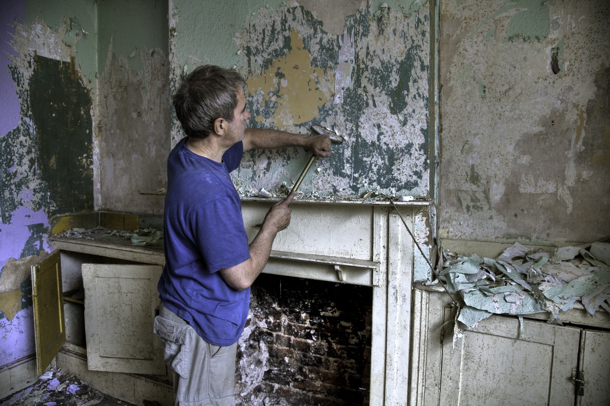 Man scraping old paint and wallpaper off a wall at the start of a house renovation project.